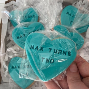 May include: Heart-shaped cookies with turquoise frosting and black text reading "MAX TURNS TWO" are packaged in clear cellophane. The cookies are arranged in a cardboard box with white paper filler.