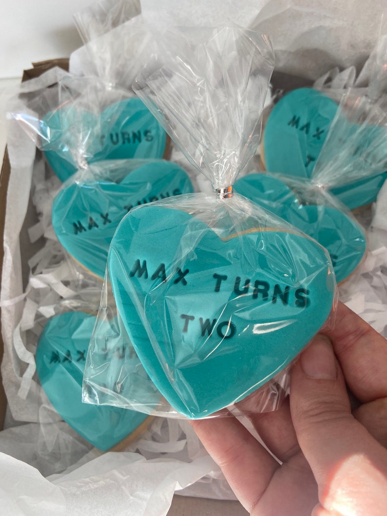 May include: Heart-shaped cookies with turquoise frosting and black text reading "MAX TURNS TWO" are packaged in clear cellophane. The cookies are arranged in a cardboard box with white paper filler.