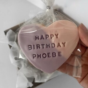 May include: A heart-shaped cookie, split diagonally with lavender and peach colors, is packaged in clear cellophane. The cookie is embossed with the words "HAPPY BIRTHDAY PHOEBE". The cellophane is tied with a silver twist tie.