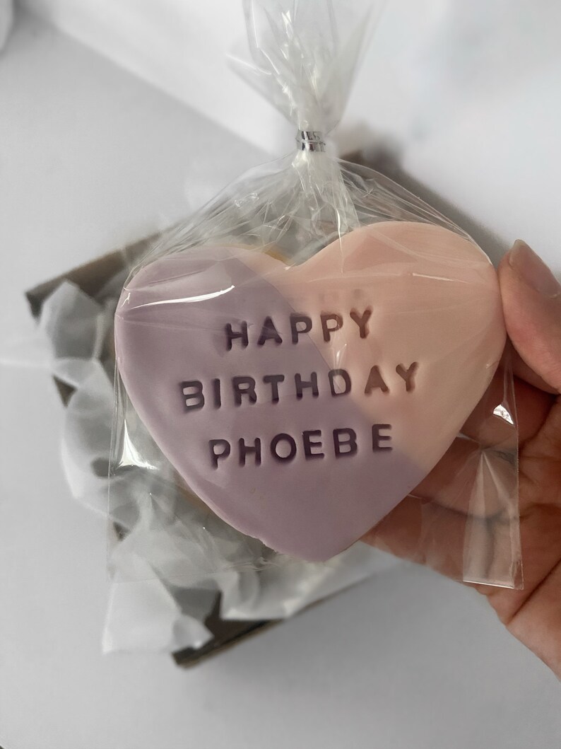 May include: A heart-shaped cookie, split diagonally with lavender and peach colors, is packaged in clear cellophane. The cookie is embossed with the words "HAPPY BIRTHDAY PHOEBE". The cellophane is tied with a silver twist tie.