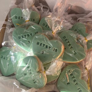 May include: A box filled with heart-shaped cookies. Each mint green cookie is individually wrapped in clear cellophane and tied with gold string. The cookies have the text "BETSY IS 2" in black.
