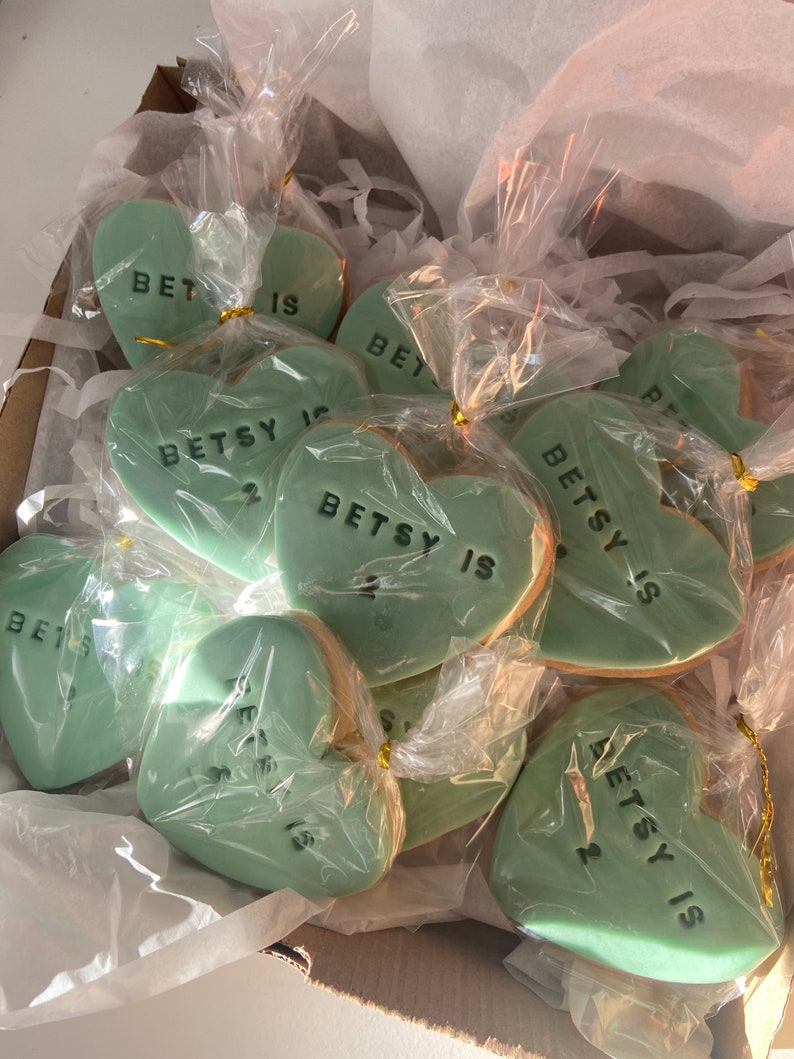 May include: A box filled with heart-shaped cookies. Each mint green cookie is individually wrapped in clear cellophane and tied with gold string. The cookies have the text "BETSY IS 2" in black.