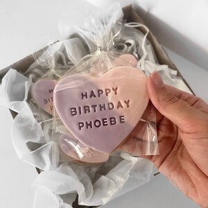 May include: Heart-shaped cookies with a two-tone pink frosting, packaged in clear cellophane and a cardboard box. The cookies are inscribed with "HAPPY BIRTHDAY PHOEBE".
