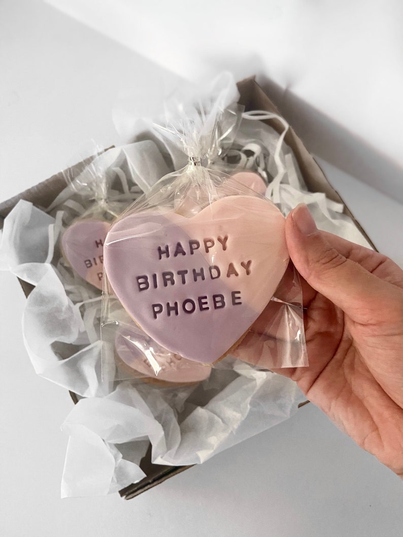 May include: Heart-shaped cookies with a two-tone pink frosting, packaged in clear cellophane and a cardboard box. The cookies are inscribed with "HAPPY BIRTHDAY PHOEBE".