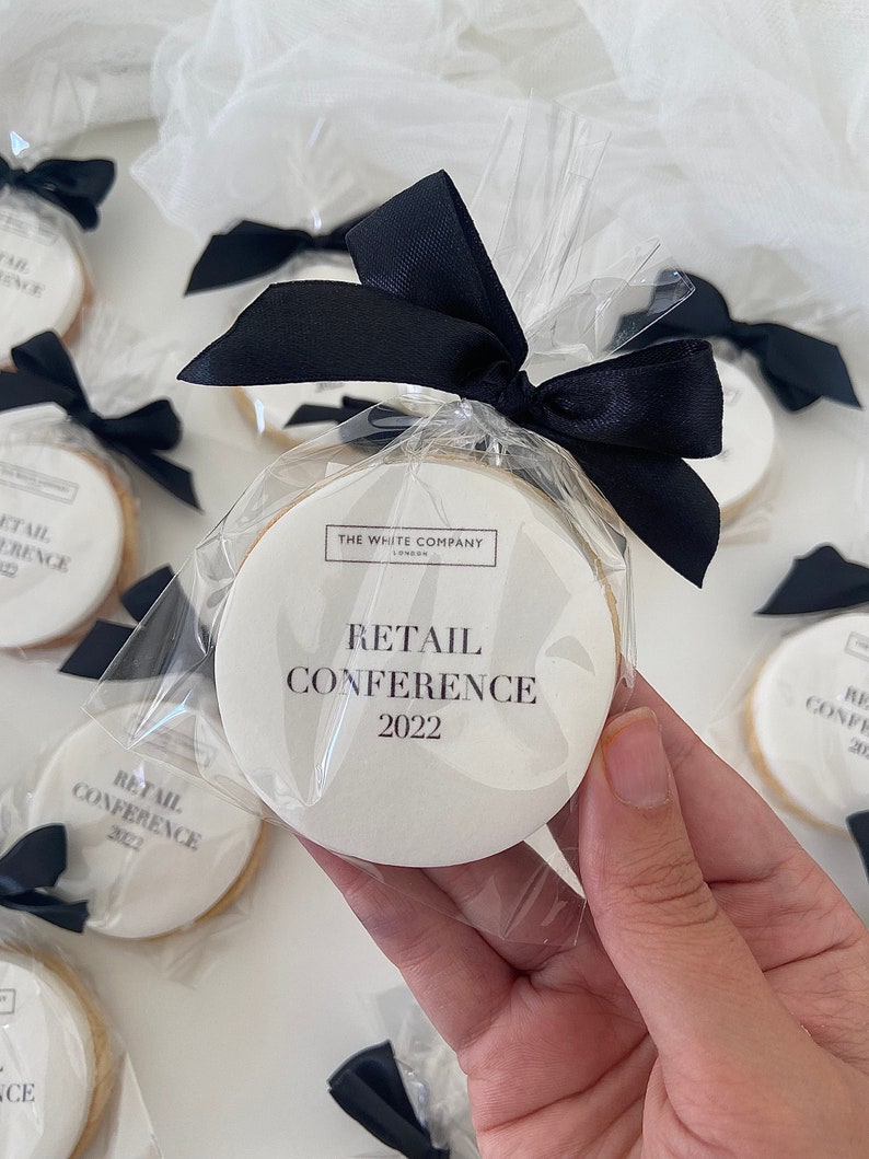May include: White round cookies with black text that reads "The White Company" and "Retail Conference 2022". The cookies are individually wrapped in clear plastic bags with black ribbon bows.