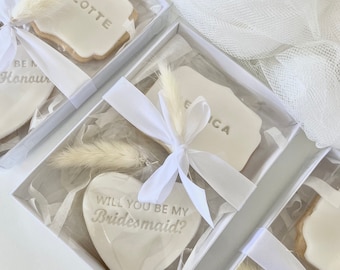 Puede incluir: Cuatro cajas blancas con tapas transparentes contienen galletas decoradas con glaseado blanco. Las galletas tienen forma de corazón y rectángulo. La galleta en forma de corazón dice "¿Quieres ser mi dama de honor?" y las galletas rectangulares tienen nombres escritos en ellas. Cada caja tiene un pequeño ramo de hierba de la pampa blanca seca.
