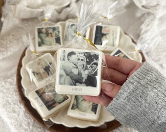 May include: A collection of square cookies with black and white photos printed on them. The cookies are individually wrapped in clear plastic bags with gold ribbon ties. The cookies feature a variety of photos, including a couple in a car, a couple standing together, and a couple holding hands. The cookies are arranged on a brown wooden surface.