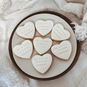 Puede incluir: Un plato de galletas en forma de coraz&oacute;n con glaseado blanco. Cada galleta tiene el texto "MR & MRS WILSON 09.03.23" en letras plateadas. Las galletas est&aacute;n dispuestas en un plato blanco con borde de madera.