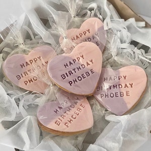 May include: Heart-shaped cookies with a two-tone pink and purple frosting, individually wrapped in clear cellophane. Each cookie is inscribed with "HAPPY BIRTHDAY PHOEBE" in embossed lettering. The cookies are nestled in a box with white tissue paper.