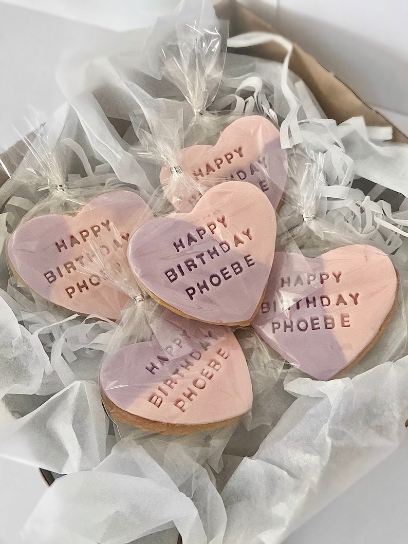 May include: Heart-shaped cookies with a two-tone pink and purple frosting, individually wrapped in clear cellophane. Each cookie is inscribed with "HAPPY BIRTHDAY PHOEBE" in embossed lettering. The cookies are nestled in a box with white tissue paper.