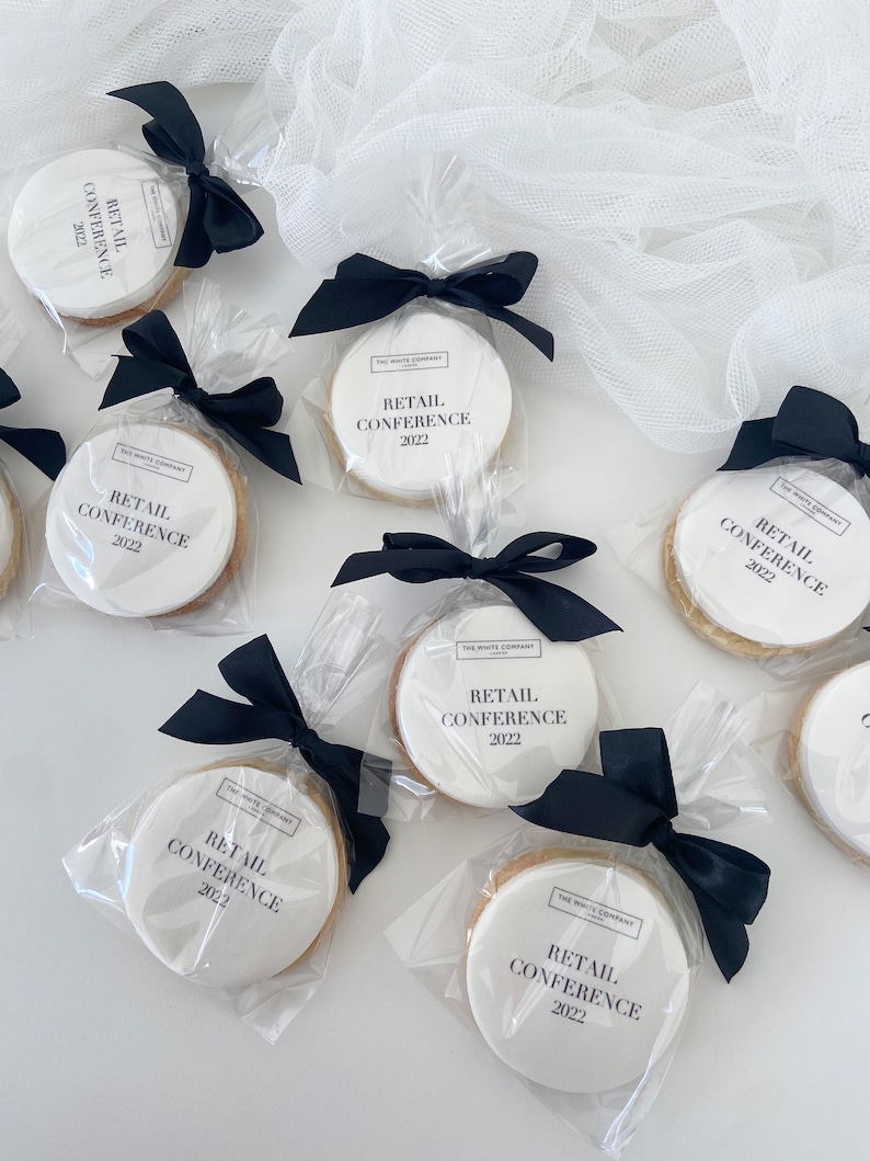 May include: White round cookies with black text that reads "The White Company" and "Retail Conference 2022" are individually wrapped in clear cellophane bags and tied with black ribbon.