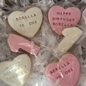 May include: A collection of heart-shaped cookies, some pink and some white, are packaged in a box. The cookies have messages like "Happy Birthday Sorella" and "Sorella is One" written on them. There is also a cookie shaped like a high-heeled shoe.