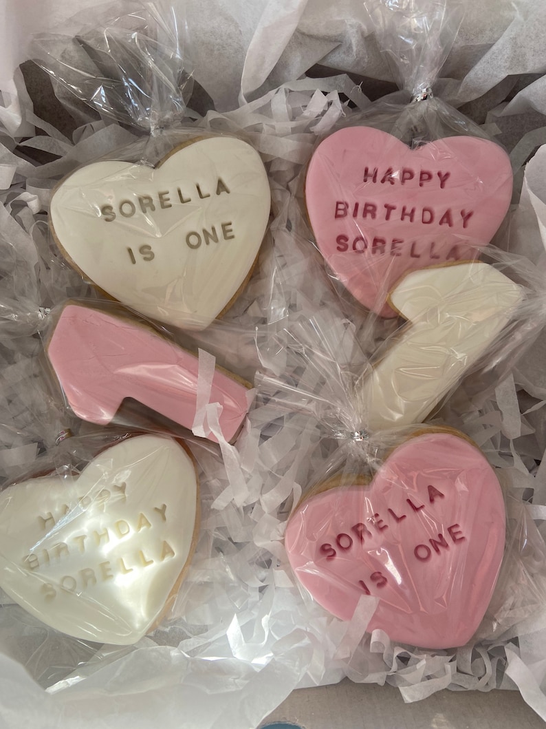 May include: A collection of heart-shaped cookies, some pink and some white, are packaged in a box. The cookies have messages like "Happy Birthday Sorella" and "Sorella is One" written on them. There is also a cookie shaped like a high-heeled shoe.