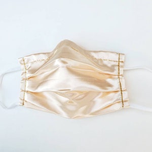 May include: A pleated face mask made of light beige satin with gold stitching and white elastic ear loops.