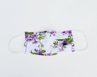 May include: A white face mask with a purple floral pattern. The mask has white elastic ear loops.