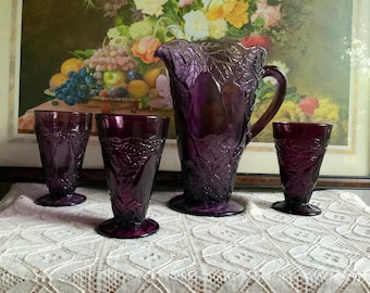 May include: A set of four purple glass pitcher and glasses with a leaf design. The pitcher has a handle and a wide mouth. The glasses are tall and slender.