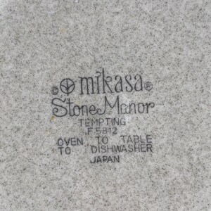 Pode incluir: Close-up de um prato de cer&acirc;mica branco com um padr&atilde;o salpicado preto e cinza. O prato tem o texto "Mikasa Stone Manor Tempting F 5812 Oven to Table Dishwasher Japan" impresso na parte inferior.