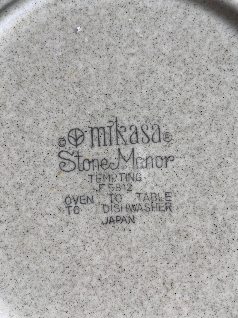 Pode incluir: Close-up de um prato de cer&acirc;mica branco com um padr&atilde;o salpicado preto e cinza. O prato tem o texto "Mikasa Stone Manor Tempting F 5812 Oven to Table Dishwasher Japan" impresso na parte inferior.