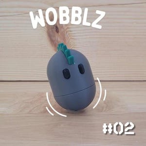 May include: A gray, egg-shaped toy with a green fin on top and two black oval eyes. The word "WOBBLZ" is above the toy, and the number "#02" is below it. The toy is on a light brown wooden surface.