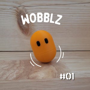 Puede incluir: Un juguete naranja en forma de huevo con dos ojos negros y la palabra "WOBBLZ" encima. El juguete está ligeramente inclinado, con líneas curvas que sugieren movimiento. El número "#01" está debajo. El fondo es una superficie de madera marrón claro.
