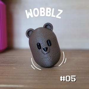 May include: A brown bear-shaped toy with the word "WOBBLZ" above it and the number "#05" below. The toy has black eyes and a small nose. The bear is on a wooden surface.
