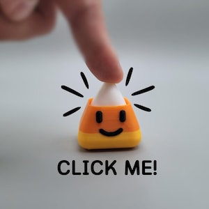 Puede incluir: Un caramelo de maíz con una cara sonriente, en naranja, amarillo y blanco, con el texto "CLICK ME!". Un dedo toca la punta blanca del caramelo de maíz, con líneas negras que irradian hacia afuera.