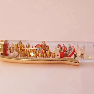 May include: A white pen with a gold cap and a clear barrel filled with gold beads, stars, and rainbow-colored shapes.