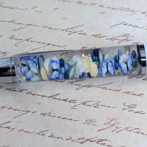 May include: A clear pen with a white barrel and a silver cap. The pen is filled with blue, white, and green flowers and butterflies. The pen is lying on a piece of paper with handwritten text.
