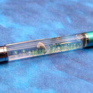 May include: A clear pen with a silver cap and a turquoise body. The pen contains green and white beads that move when the pen is shaken.