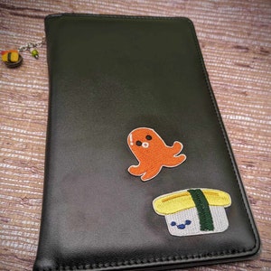 May include: Black leather notebook with a cute octopus and sushi embroidery. The octopus is orange and the sushi is yellow with green seaweed.