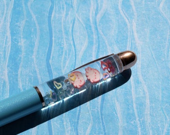 Op de afbeelding: Een lichtblauwe pen met een doorzichtige cilinder gevuld met glitter en kleine kleurrijke bedeltjes. De pen heeft een zilveren clip en een zilveren punt.