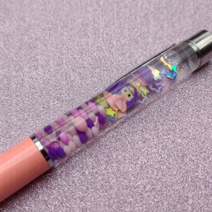 May include: A pink and clear pen with a variety of colorful beads and charms inside. The pen is on a purple sparkly background.