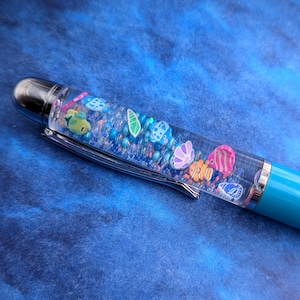 May include: A turquoise blue pen with a clear barrel filled with glitter and colorful fish and seashells. The pen is on a blue background.