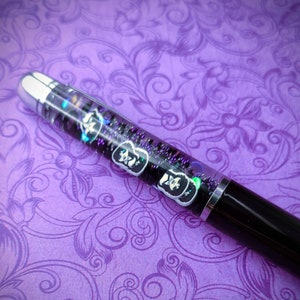 May include: A black and silver pen with a clear barrel filled with glitter and small black and white images. The pen is lying on a purple background with a floral pattern.