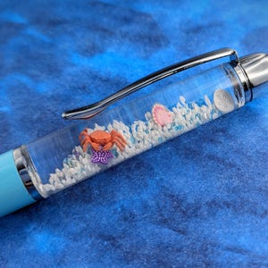 May include: A clear pen with a blue cap and a silver top. The pen contains a small crab, a pink seashell, and white sand.
