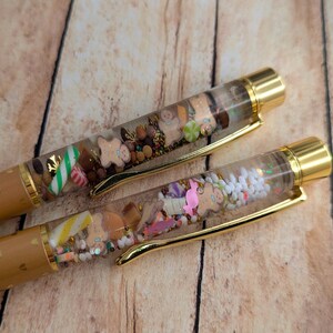 May include: Two gold pens with clear barrels filled with colorful glitter, sprinkles, and gingerbread men. The pens have brown barrels with gold hearts.
