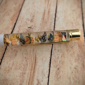 May include: A clear pen with a white cap and gold accents. The pen is filled with small, colorful objects, including bees, honeycomb, and leaves.