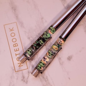 May include: Two silver pens with green and black alien faces on the barrel, lying on a white marble notebook with the word "NOTEBOOK" printed in gold.