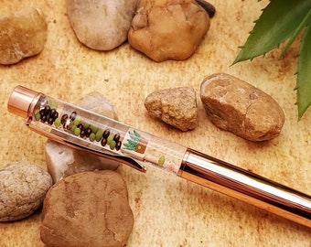 May include: A rose gold pen with a clear barrel filled with green and brown glitter. The pen is lying on a brown surface with small rocks.