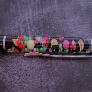 Puede incluir: Un bolígrafo negro con un barril transparente lleno de caramelos de colores. El bolígrafo tiene un clip plateado y una tapa plateada.