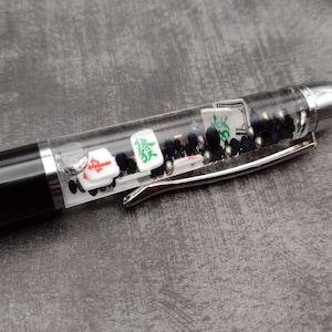 May include: A black and silver pen with a clear barrel filled with black beads and mahjong tiles. The tiles have Chinese characters and red and green accents.