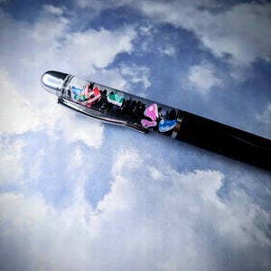 May include: A black pen with a clear section filled with colorful miniature sneakers. The pen is lying on a white and blue cloudy background.