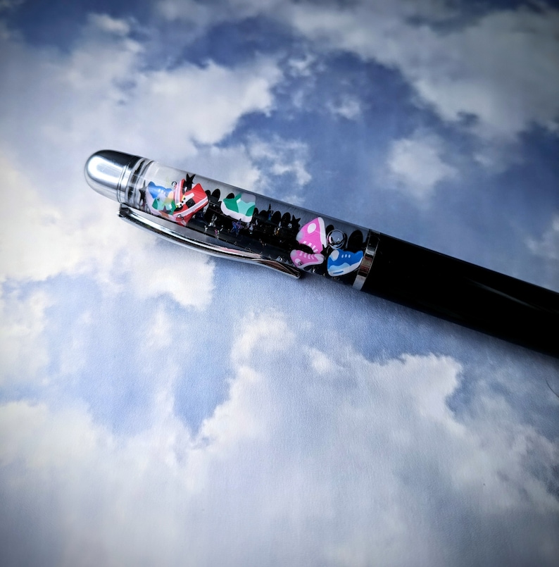 Op de afbeelding: Een zwarte pen met een doorzichtig gedeelte gevuld met kleurrijke miniatuur sneakers. De pen ligt op een witte en blauwe wolkachtige achtergrond.