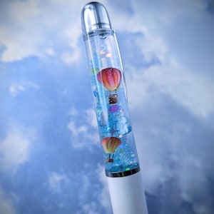 May include: A clear pen with a silver top and a white bottom. The pen is filled with blue liquid and contains small, colorful hot air balloons.