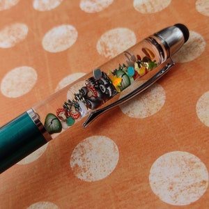 May include: A teal and silver pen with a clear barrel filled with miniature woodland-themed decorations. The pen features a black stylus tip and a silver clip. The decorations include a raccoon, mushrooms, and colorful gems.
