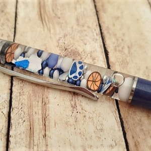 May include: A blue and silver pen with a clear section filled with colorful beads and a basketball design.
