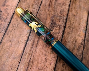 May include: A ballpoint pen with a dark teal lower barrel and a clear upper barrel filled with small decorative items including a gold-colored Bigfoot silhouette, miniature pinecones, and various green, brown, and blue beads with gold trim.