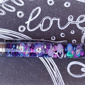 May include: A purple pen with a clear barrel filled with colorful beads and charms. The pen is lying on a black surface with white doodle art.