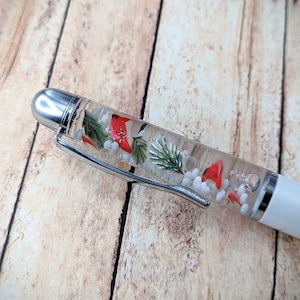 May include: A clear pen with a white cap and a silver clip. The pen contains red cardinals, green pine branches, white glitter, and white beads.