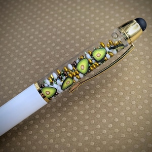 May include: A white pen with a clear barrel filled with gold and white beads and avocado slices. The pen has a gold top with a stylus tip.